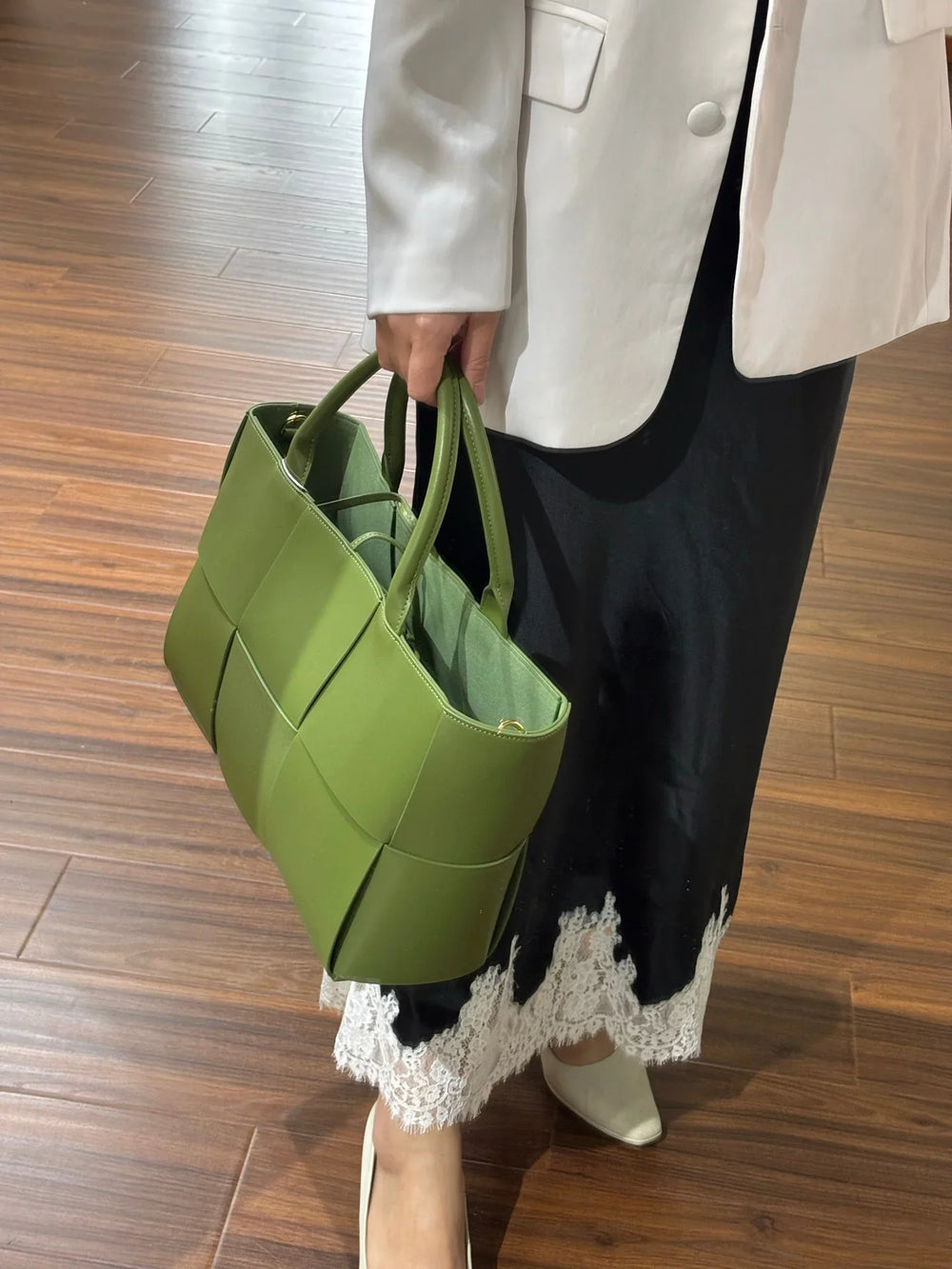 Woman holding a green woven leather tote bag with top handles, elegant outfit, wood floor