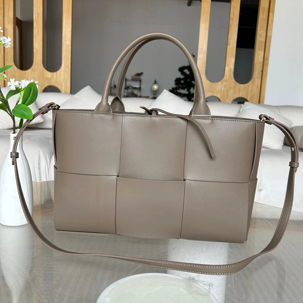 Large taupe woven leather tote bag with top handles and shoulder strap on a glass table