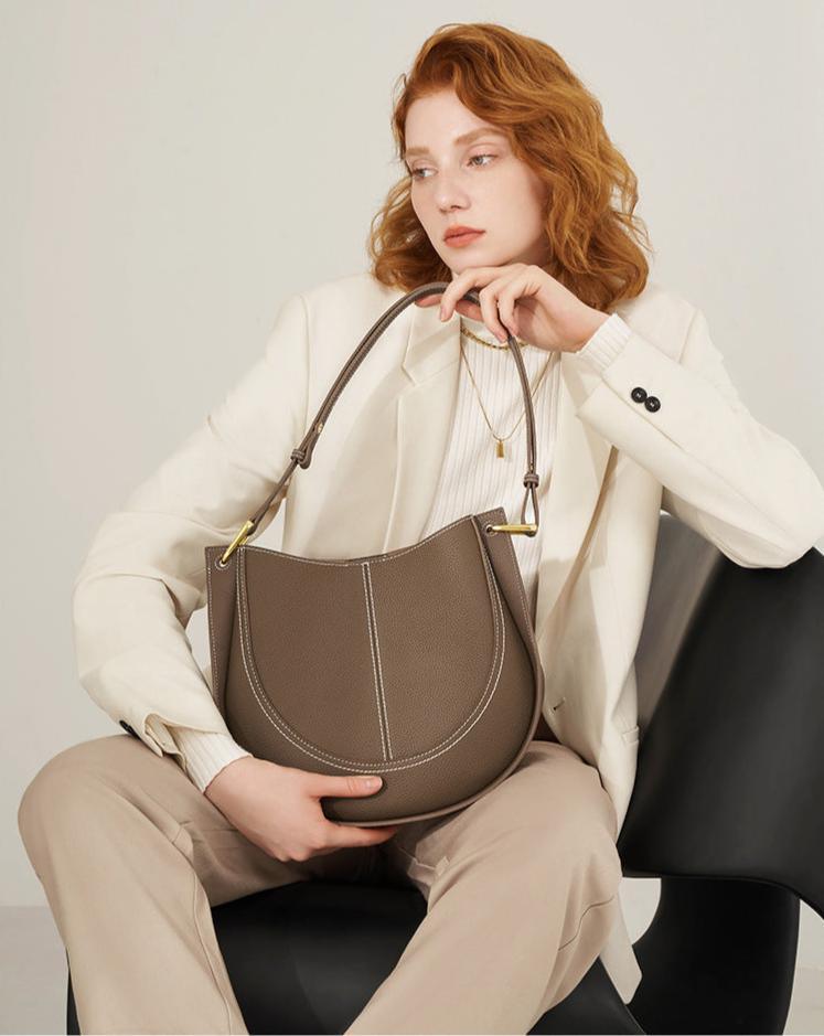 Woman in cream suit holding taupe smooth calfskin leather saddle bag with white stitching