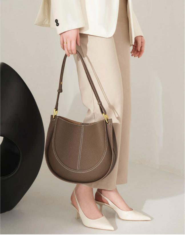 Woman holding smooth brown calfskin leather saddle bag with gold hardware, wearing cream suit and heels