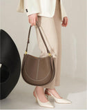 Woman holding smooth brown calfskin leather saddle bag with gold hardware, wearing cream suit and heels