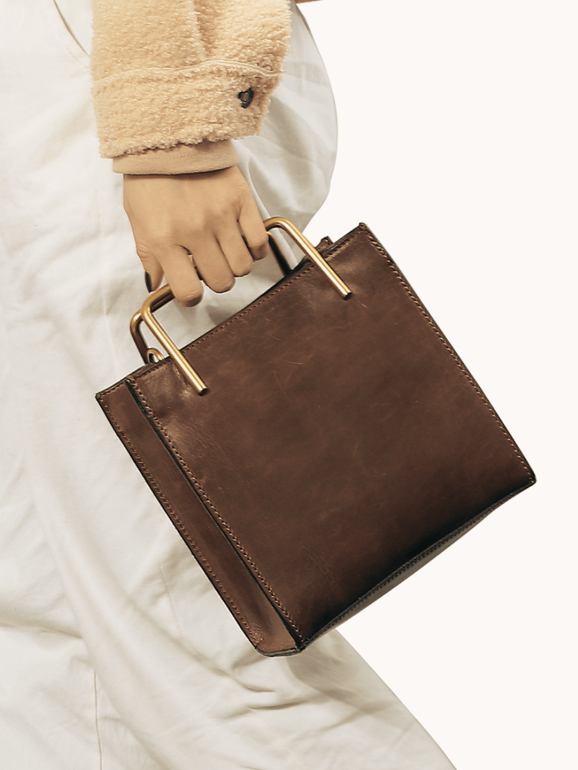 Woman holding a brown leather handbag with gold handles, stylish and minimalist design
