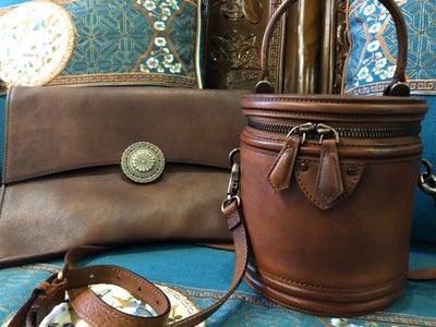 Handcrafted brown leather clutch with decorative metal accent and round leather shoulder bag on patterned fabric background.