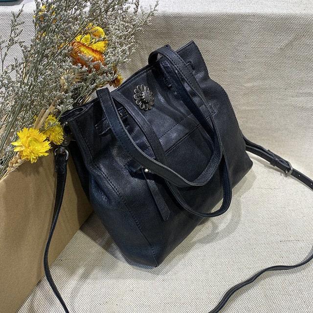 Black leather women's shoulder bag with flower charm, displayed beside dried flowers
