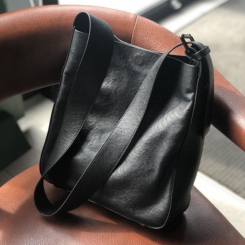Black full grain leather shoulder bag with wide strap on a brown chair