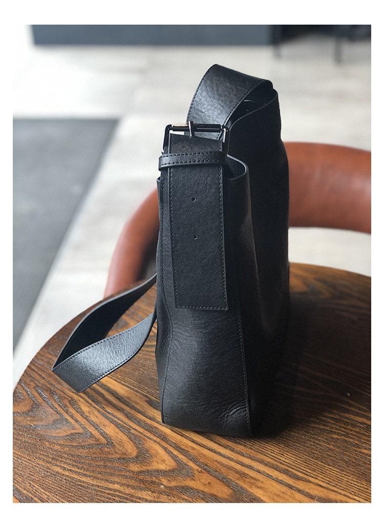 Black full grain leather shoulder bag with wide strap on a wooden table, crossbody style