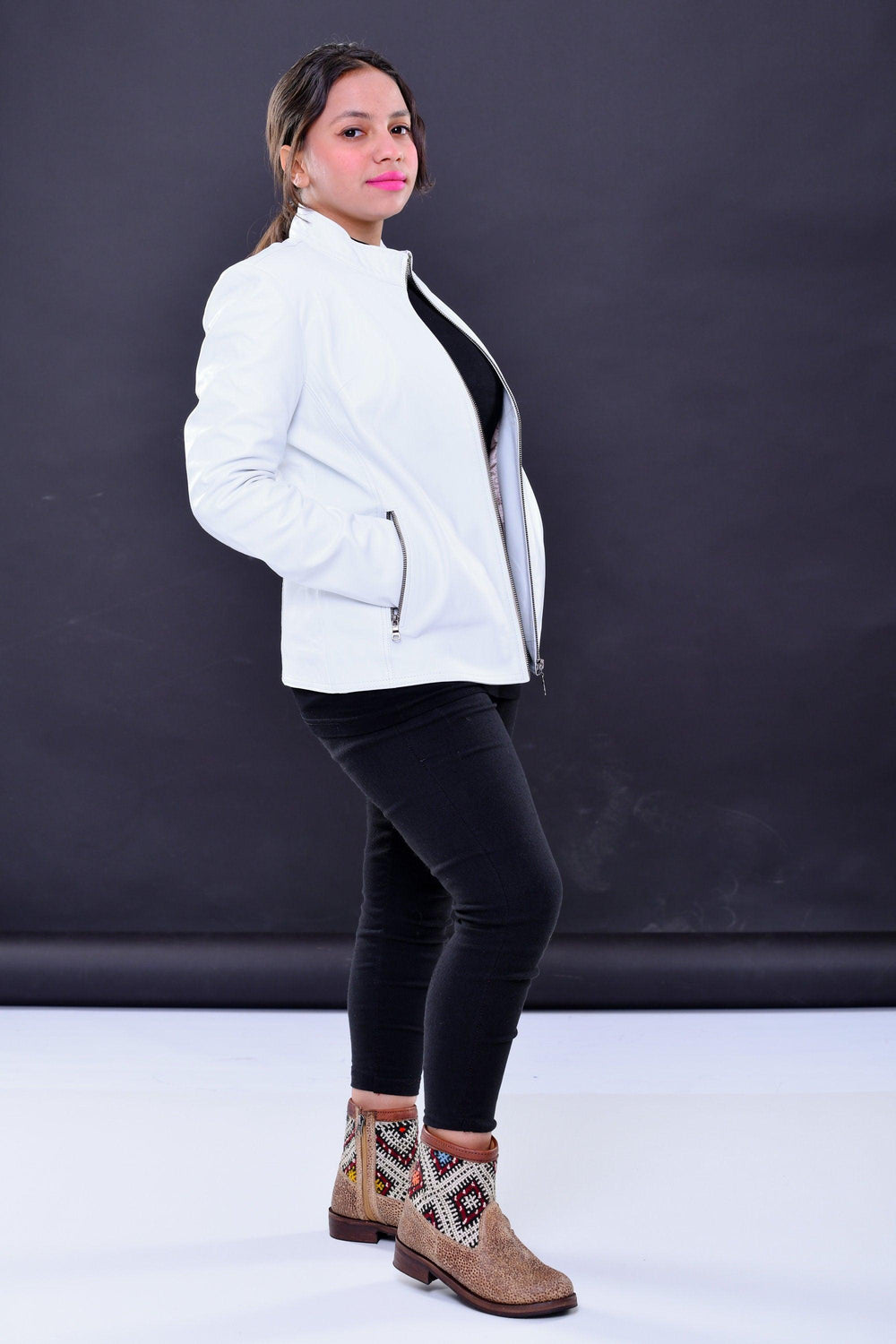 Woman wearing white genuine leather biker jacket with zipper pockets, black pants, and tan patterned boots, posing against dark background.