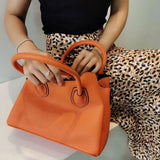 Woman holding a textured orange leather handbag with double handles, wearing a leopard print skirt.