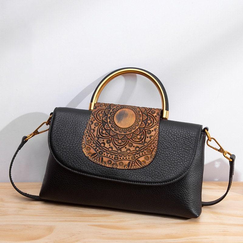 Black leather handbag with embossed brown mandala flap and gold metal handle on wood surface