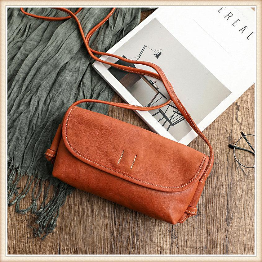 Brown cowhide leather shoulder bag with long strap on wooden surface, next to magazine and scarf