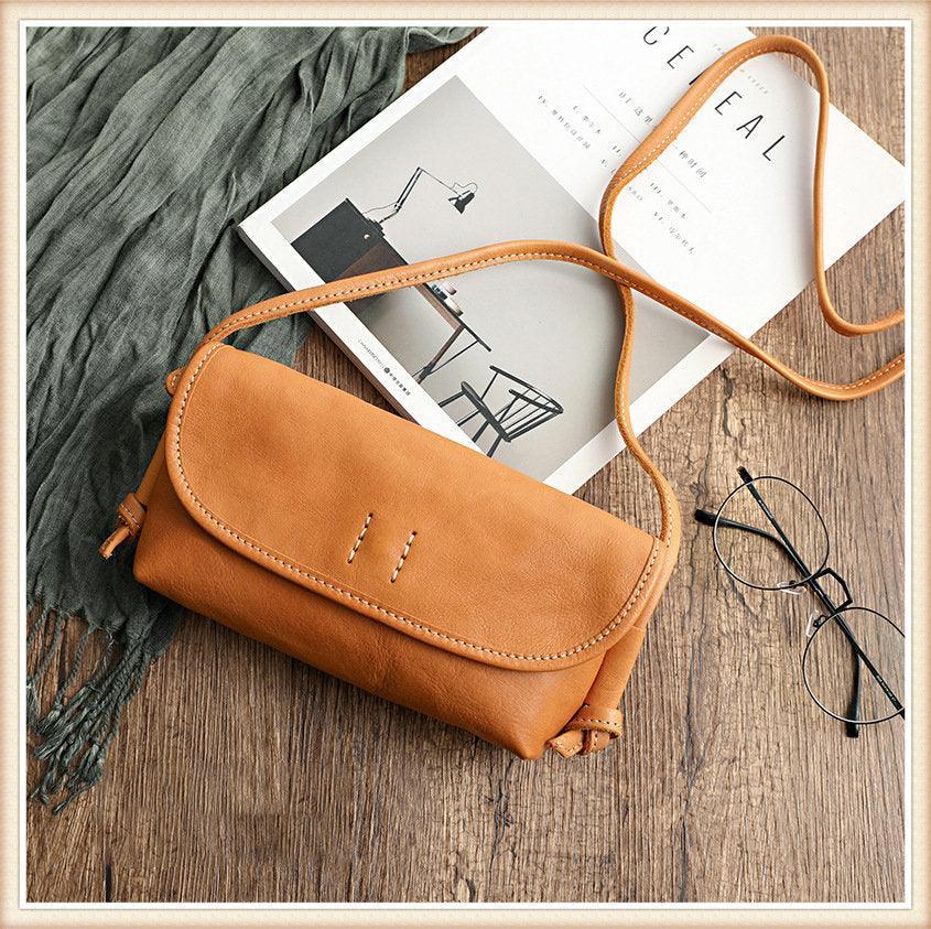 Tan cowhide leather shoulder bag with flap closure, displayed on wooden surface beside glasses and magazine.
