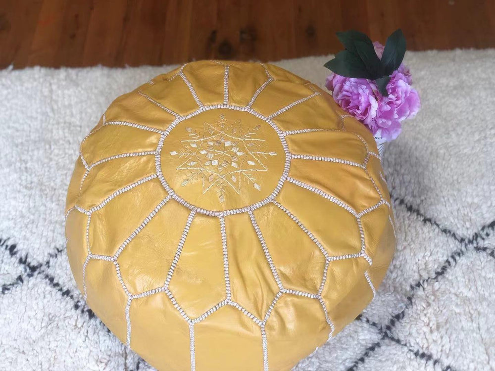 Handmade light yellow Moroccan leather pouf footstool on a textured rug, vase with pink flowers nearby