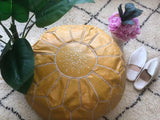 Handmade light yellow leather Moroccan pouf on patterned rug with slippers, flowers, and plant