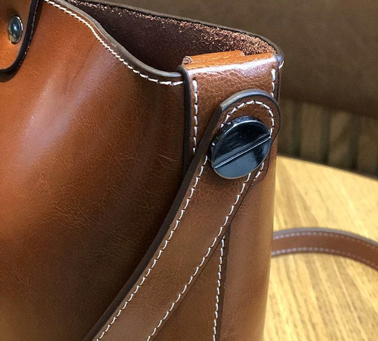 Close-up of brown leather bucket bag with white stitching and crossbody strap on wood table
