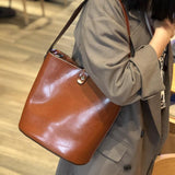 Brown leather bucket shoulder bag worn crossbody by woman in neutral coat indoors
