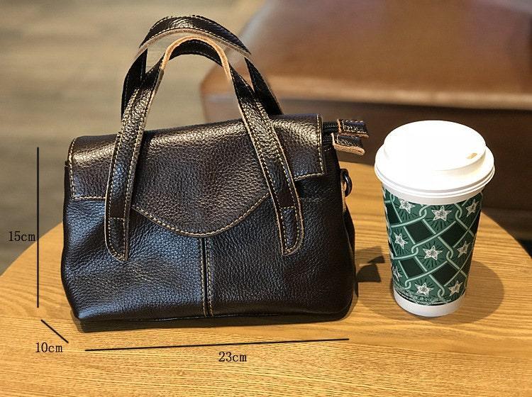 Small black leather crossbody handbag with top handles, shown on a table beside a coffee cup, size 23x15x10cm.