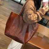 Large brown minimalist leather tote bag with double shoulder straps, worn by a woman indoors