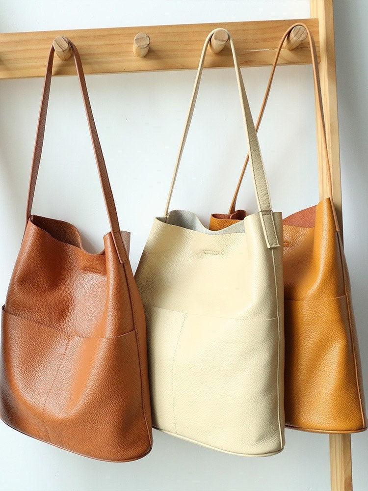 Three handcrafted leather tote bags with front pockets in tan, cream, and caramel, hanging on a wooden rack.