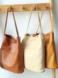 Three handcrafted leather tote bags with front pockets in tan, cream, and caramel, hanging on a wooden rack.